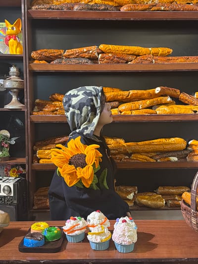 A person wearing a camouflage hat and a black top with a large sunflower design is partially visible in front of a wooden shelf filled with artificial bread loaves. The shelf contains various other items including a yellow rabbit toy, vintage machinery, and floral arrangements. A wooden tray displays artificial cupcakes and donuts. The location is Thuận Hóa Ward, Vietnam.
