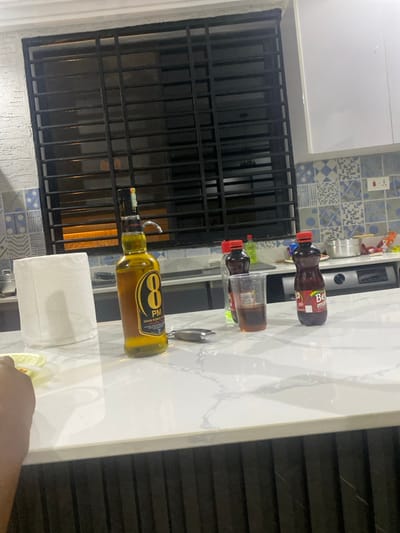 The image captures a kitchen counter scene in a residential setting, likely in Adenta Municipality, Ghana, given the context. The prominent feature is a white, marble-patterned countertop holding various items.
On the counter, there are several drink bottles: a brown plastic bottle labeled "Beri Mornu," and another similar bottle next to a clear plastic cup containing a dark beverage. A larger, yellow-liquid glass bottle, identifiable as "8 PM" with the text "CABIN BLENDED" on its label, stands near a silver bar tool or bottle opener. To the left of the "8 PM" bottle, a roll of white paper towels is placed. In the bottom right corner, a human hand is partially visible, holding a small, yellow, possibly plastic item or snack.
The background features a window secured with a black metal grille or security bars, offering a dark, indistinct view of the outside. Above the counter and to the left of the window, there's a white cabinet and a backsplash adorned with blue and white patterned tiles. Other kitchen appliances, such as a blender, and metal pots are faintly visible on an upper shelf or counter. The overall scene suggests a casual indoor environment, possibly during drink preparation or a small gathering.