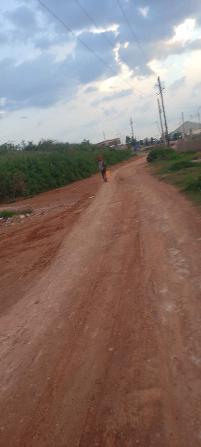 In Rayfield, Nigeria, a dirt road cuts through a semi-arid landscape under a cloudy sky. Lush green vegetation lines the left side of the road, interspersed with patches of bare, reddish-brown earth and scattered debris. On the right, more sparse vegetation and the beginnings of construction are visible, including utility poles and what appear to be small buildings. A lone figure, carrying a yellow container on their head, walks away from the camera down the center of the road, dressed in dark clothing and a red head covering. The road itself is rutted and dusty, showing signs of wear from vehicular traffic. The overall impression is one of a developing area, likely on the outskirts of a town or city, where daily life involves the transport of water.
