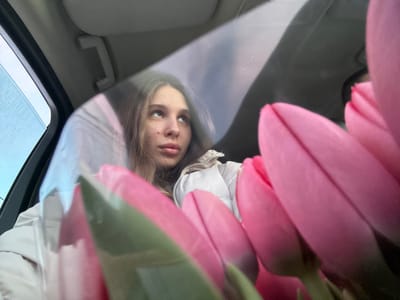 The image depicts the interior of a vehicle in Slavyansk-na-Kubani, Russia. A young female individual is situated in the left-center of the frame, facing slightly rightward with her gaze directed upwards. She has long, light brown hair and is wearing a light-colored, possibly white or beige, quilted jacket. A small dark mark or mole is visible on her right cheek. In the foreground, occupying the right half of the image, is a bouquet of multiple pink tulip buds, tightly closed and displaying some green stem and leaf elements. The tulips appear to be wrapped in a transparent material, causing reflections across their surface. The vehicle's interior shows a light-colored grab handle and part of the headliner in the upper sections. A dark vertical element, consistent with a car's pillar or door frame, is present on the far left. Through a window in the upper-left, a blurred outdoor environment with blue and grey tones is visible, suggesting an overcast sky. The scene is illuminated by ambient daylight.