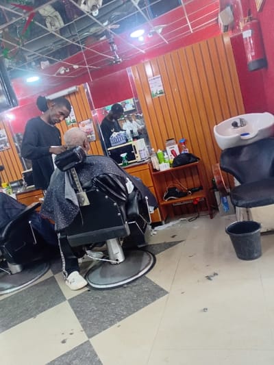 The scene is set inside a barbershop or hair salon in Dorayi, Nigeria. A male barber, wearing a black long-sleeved shirt, stands behind a seated client, actively trimming the client's short, light-colored hair with clippers. The client is draped in a black and white polka-dotted cutting cape and occupies a black barber chair with a chrome base. The barbershop interior features red-painted walls with lower sections of vertical wooden paneling. The ceiling consists of reflective panels, from which light fixtures and a ceiling fan are suspended. The floor is tiled in a checkerboard pattern of light grey and dark grey squares. To the right, a white ceramic hair wash basin is mounted, accompanied by a black chair, and a black plastic bucket rests on the floor beneath it. A red fire extinguisher is mounted on the red wall in the upper right. Mirrors cover a significant portion of the back wall, reflecting the barber, the interior, and a television screen displaying what appears to be individuals. Various hair care products and tools are arranged on a wooden shelving unit and countertop behind the barber and client. A second black barber chair is partially visible on the far left.