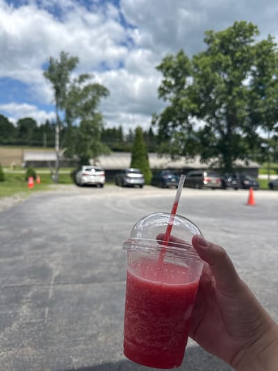 A first-person perspective shot shows a hand holding a clear plastic cup filled with a pinkish-red frozen beverage. A red and white striped straw protrudes from the cup, and the drink appears to be slushy or blended. The background is blurred but suggests an outdoor setting. Trees with green foliage are visible, and the sky is partly cloudy with blue patches. The ground appears to be asphalt or concrete. Further in the background, several cars are parked in a line, and orange traffic cones are interspersed among them. A few buildings with light-colored siding are also visible. The scene is shot during the day, likely late morning or afternoon, given the bright but diffused light. The overall impression is casual, perhaps a stop at a roadside stand or a casual outing on a warm day. There is no visible text in the image.