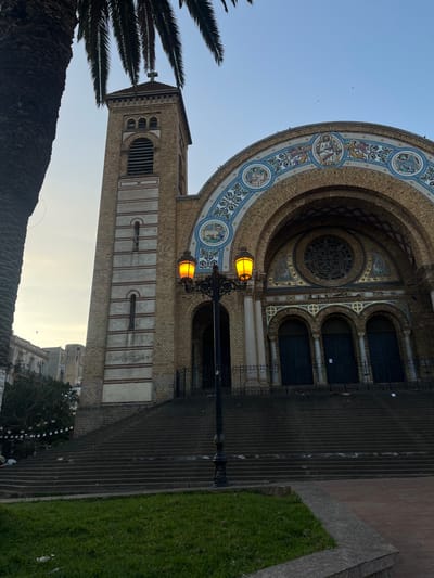 The image displays the exterior of the Sacré-Cœur Cathedral, located on Place du 1er Novembre in Oran, Algeria. The central subject is a large, multi-story building constructed primarily of light-brown brick, featuring a prominent bell tower on the left topped with a cross. The tower includes vertical white banding and arched belfry openings. The main facade presents a grand central arch adorned with extensive mosaic artwork in blue, gold, and white, depicting religious figures and intricate patterns, positioned above a large circular rose window. Beneath the rose window are three dark, arched entrance doors, fronted by dark metal railings. A wide, multi-tiered stone staircase leads up to the building's entrance from the foreground. Centered in the mid-ground, a black, ornate streetlamp with two illuminated orange-yellow globes partially obscures the lower facade and stairs. A substantial palm tree trunk and fronds frame the scene from the far left. In the immediate foreground, a patch of green grass is visible within a raised curb. Distant urban buildings with balconies and string decorations are partially visible in the background to the left. The sky is clear and light blue, suggesting a dawn or dusk setting. No individuals or discernible activity are present within the frame.
