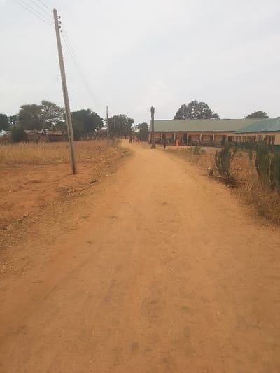The image shows a dirt road in Kwal, Nigeria (latitude: 9.805, longitude: 8.647). The road is brown and dry and leads toward a large, yellow building with a green roof. The building appears to be a school. It is situated on the right side of the image, and there are a few trees and shrubs in front of it. On the left side of the image, there is a wooden utility pole with wires running to another pole in the distance. Several people are walking on the road in the distance. There are a few small trees and bushes along the road, and the sky is gray and overcast. The environment appears to be rural and dry. The image was likely captured with a smartphone, based on the image quality. The camera is positioned at ground level, facing forward on the dirt road. The image is clear and bright enough for forensic examination.