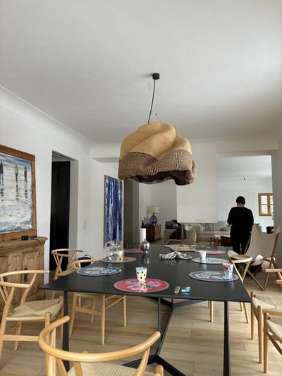 The image shows a dining room in a house located at latitude 46.514, longitude 9.859 in Celerina/Schlarigna, Switzerland. There is a large, black rectangular table with six chairs surrounding it. The table is set with placemats and cups. A large, woven, light brown, and dark brown lamp is hanging from the ceiling.  The room has light walls and a wooden floor. A woman stands in the background to the right of the table, wearing a dark sweater and dark pants, and she is looking towards a doorway in the right corner of the image. The doorway is blocked by a white lounge chair.  A painting on the wall to the left of the dining table is partially visible. There is a wooden cabinet to the left of the table.  The scene is a still life shot of a dining room.  The image appears to have been taken during the day.