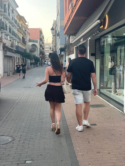 In La Línea De La Concepción, Spain, a couple walks away from the camera on a brick-paved street. The woman, on the left, wears a dark crop top and a dark, short, ruffled skirt, with light-colored platform sandals. She carries a light-colored shoulder bag and has dark hair. The man, on the right, wears a black t-shirt and light-colored denim shorts with white sneakers. They are walking on the sidewalk adjacent to shops. The street features buildings with balconies on the left and shopfronts with awnings on the right. Several other pedestrians are visible in the background, as well as trees lining the sidewalk. The sky is overcast and light.