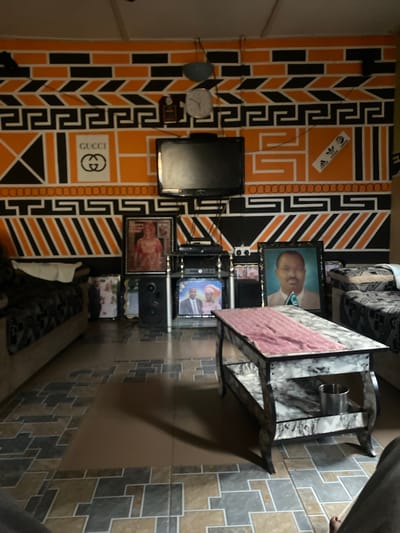 This image depicts the interior of a domestic living space located in Uffah, Nigeria. The room features walls extensively decorated with geometric patterns in black and orange, outlined with white lines. Distinct patterns include horizontal bricks, chevrons, a Greek key design, and various stripes and circles. A white "GUCCI" logo with interlocking Gs and an Adidas logo sticker are visible on the orange sections of the wall. A round wall clock is mounted on the upper left wall.
A black television set is positioned on a multi-tiered black entertainment stand in the center. Below the television, electronic components and at least one large black speaker are arranged. Numerous framed photographs are displayed on the floor around the entertainment stand, depicting individuals and couples. A prominent framed portrait to the right features a man in a suit and tie.
The room contains two upholstered sofas, partially visible on the left and right, with patterned fabric and light-colored bases. A white cloth is draped over the armrest of the sofa on the left. In the foreground, a rectangular coffee table with a faux marble finish and a lower shelf holds a pink fabric runner. A silver metallic cup rests on the table's lower shelf. The floor is tiled with a mosaic-like pattern in shades of brown, gray, and beige. A human leg and bare foot are visible in the lower right corner, indicating a person is seated out of frame.