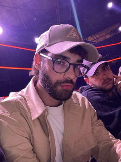Two men pose for a selfie. The main subject wears a light-colored cap with decorative stitching, round glasses, a white t-shirt, and a tan jacket. He has a dark beard and is closer to the camera. Behind him is another man wearing a white cap and a dark top. The background is a mix of purple and orange lights, indicating an indoor setting, possibly a venue with ambient lighting.