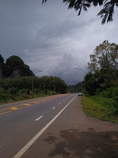 The image depicts a paved road in Khao Khram, Thailand, flanked by lush greenery and dramatic karst formations under an overcast sky. The road features yellow markings on the left and a solid white line on the right. A dark-colored vehicle is parked on the right shoulder. Streetlights line the road. The foreground shows the pavement with shadows.
