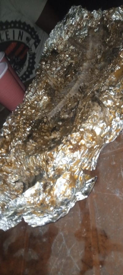A spent fish skeleton sits on a foil-lined surface, remnants of the meal visible. Beside it, a red plastic cup rests near the edge of what appears to be a t-shirt displaying a logo or graphic, slightly obscured. The overall scene is dimly lit and somewhat cluttered, suggesting a casual setting. A dark wooden table underlies the aluminum foil.