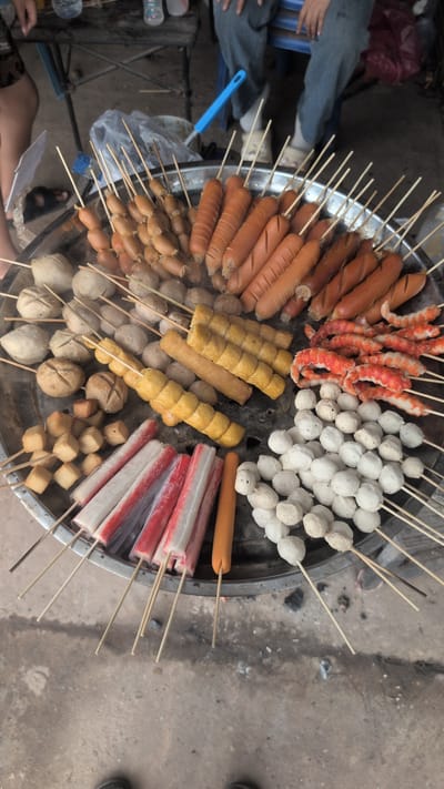The image shows a close-up, overhead view of a large metal tray filled with various skewers of food, likely being prepared for sale. The food items include sausages, meatballs, imitation crab sticks, and what appear to be fried fish cakes or similar items. Some of the food is already cooked and steaming. In the background, partially visible, are people sitting on chairs and a table with some items on it. The ground appears to be concrete and dirt, with some scattered debris. The scene is consistent with a street food vendor setup, possibly in a market or public area within the city of Phao, Laos, given the presence of what seems to be local people and the overall atmosphere of an outdoor food stall.