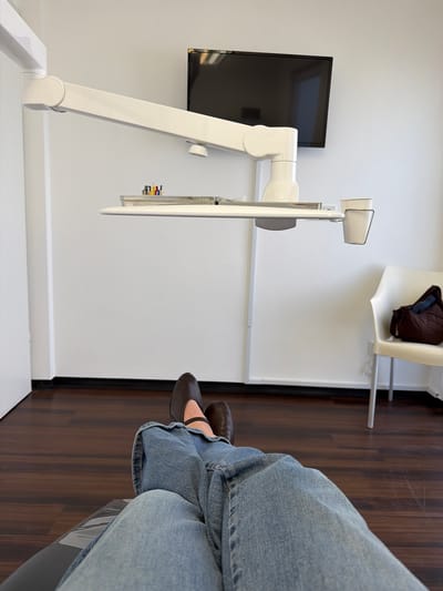 A person is seated in what appears to be a dental or medical examination room in Monsheim, Germany. The person is wearing blue jeans and brown shoes. A white examination light fixture is visible on the left. A black television screen hangs on the wall in the background. A white chair with a brown jacket on it sits in the upper background, against the wall and next to the door frame. The floor is made of dark wood.