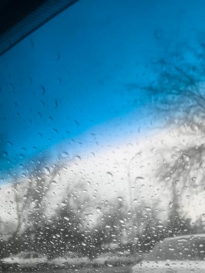 The scene depicts an exterior view through a rain-covered vehicle windshield, located in Pakhtakor, Uzbekistan. The foreground is dominated by numerous clear water droplets of varying sizes adhering to the glass, indicating precipitation or heavy condensation. Above the droplets, a dark, textured band, consistent with a vehicle's upper interior or sun visor, is visible at the top left. Beyond the windshield, the outdoor environment is largely out of focus and appears desaturated in shades of grey. Bare tree branches, indicative of dormant deciduous trees, are visible throughout the midground. Faint vertical structures, possibly streetlights or utility poles, are also discernible. A blurred, light-colored vehicle is partially visible in the lower-midground, positioned horizontally. The upper portion of the view reveals a gradient sky, transitioning from a distinct blue at the top to a lighter, overcast greyish-white near the horizon. No people, specific actions, or direct interactions are visible.