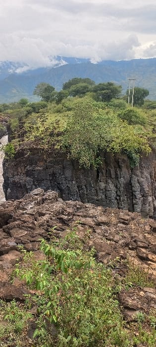 This image depicts a natural landscape in Ainamoj Village, Kenya. The foreground features rugged, dark brown rocky terrain with sparse green foliage, including a prominent green bush in the lower center. In the midground, a dark, columnar rock cliff face is visible, with a narrow body of water running through a channel to its left. The top of this cliff is covered in green grass and dense brush. The background consists of distant green, mountainous terrain partially obscured by low-hanging grey clouds. An overcast sky dominates the upper portion of the image. A utility pole structure with visible lines stands on a higher elevation in the mid-right, situated between the immediate landscape and the distant mountains. No people or animals are present.
