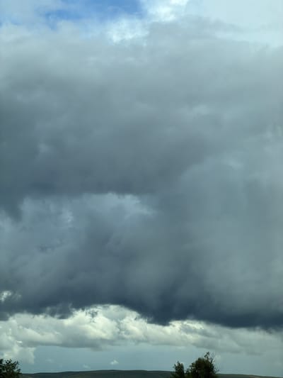 The image shows a cloudy sky over a hilly landscape. The clouds are dark and heavy, suggesting an approaching storm. There are a few trees visible in the foreground, silhouetted against the lighter sky. The landscape appears to be rural, with rolling hills covered in vegetation. The city of Верхнеозёрное, Russia is not visible in this image, as it depicts a natural landscape.