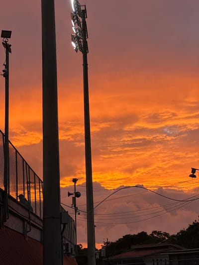 The image depicts an outdoor scene in San Antonio, Costa Rica, at dusk or dawn, characterized by a predominantly orange and red sky with layered cloud formations.

In the foreground and mid-ground, a sports or recreational facility is evident. On the far left, a section of a chain-link fence, likely part of a stadium or field enclosure, extends diagonally upwards from a structure with a reddish, corrugated metal roof. Behind the fence, several silhouetted human figures are visible, consistent with spectators or personnel.

Three prominent vertical poles are present. A dark, tall pole on the far left features a small light fixture mounted near its top. A thicker, dark pole occupies the central foreground, acting as a strong vertical element. To its right stands a tall light tower equipped with multiple active, bright floodlights, generating visible light flares. Further to the right and lower, a thinner pole supports two dark, horn-shaped speakers facing downwards and a small sensor or camera.

Multiple dark utility lines or cables traverse the mid-ground horizontally. On the far right, a partial view of another pole with a downward-facing street light fixture is visible. The lower right portion of the image reveals the dark silhouettes of buildings with discernible rooflines and dense tree canopy against the illuminated sky.