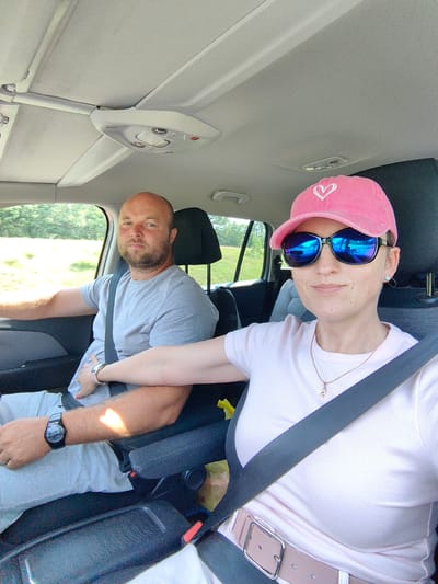 Two individuals, a male driver and a female passenger, are seated inside a car. The male driver, with a bald head and beard, wears a grey t-shirt and is looking forward. He has a watch on his left wrist. The female passenger, wearing a pink baseball cap and sunglasses, faces the camera. She has on a pink t-shirt and a necklace. A seatbelt is visible across her chest. Her arm is resting on the driver's arm. The car interior, including the ceiling and seats, is visible. Outside the car, trees and foliage can be seen, suggesting a rural or suburban setting. The image appears to be a selfie taken from the passenger's perspective. The location is identified as Dzyarzhynsk, Belarus.