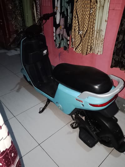 A light blue electric scooter is parked on a tiled floor indoors. The scooter has a black seat, black handlebars, and a silver handle at the rear. A small sticker with a logo is visible on the front of the scooter. The scooter is upright, supported by a kickstand. In the background, a pink wall is adorned with hanging fabrics featuring floral and abstract patterns. The lighting suggests an indoor setting, possibly a home or garage, in Sendangrejo, Indonesia.