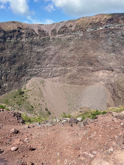 The image shows the interior of a volcanic crater, likely Mount Vesuvius, located near Ercolano, Italy. The crater walls are composed of layered rock in shades of brown and gray, with evidence of erosion and rockfalls. A large, sloping area of loose scree and gravel dominates the lower part of the crater, with sparse green vegetation growing on some slopes. The foreground consists of reddish-brown volcanic soil and rocks, suggesting the image was taken from the crater rim looking downwards into the caldera. The sky above is blue with scattered white clouds. There are no people or discernible man-made structures visible in the immediate scene.