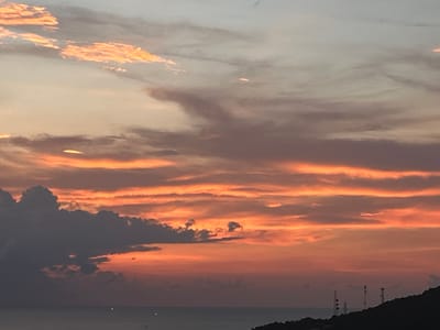 The image depicts a sunset scene over the ocean near Baan Hin Kong, Thailand. The sky is filled with layers of clouds in varying shades of orange, pink, gray, and white. Some clouds are dark and dense, while others are light and wispy. Below the clouds, the ocean is visible as a dark expanse with a few faint light spots reflecting on the surface. In the lower right corner, there is a silhouette of land with several antenna towers.