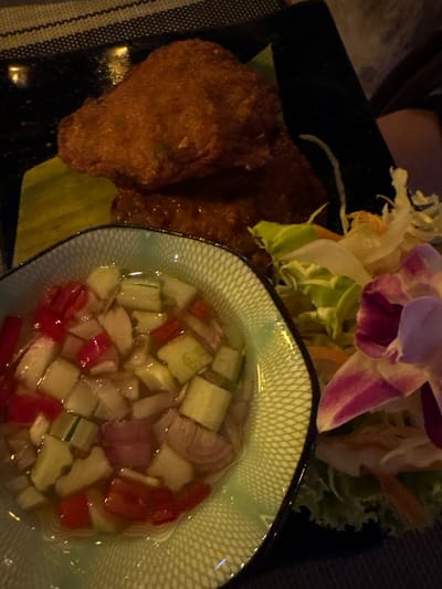 An overhead view depicts a plated meal situated on a dark, woven placemat within a dining environment in Ban Ko Li Pe, Thailand. The primary subject is a black rectangular dish containing three golden-brown, deep-fried patties, positioned on a light green leaf segment. Adjacent to this, a small, light green ceramic bowl with an embossed geometric pattern holds a clear liquid relish composed of diced red peppers, white onions, and green cucumbers. To the right of the main dish, a garnish of shredded green lettuce, pale green cabbage, and orange julienned vegetables is present, topped with a single purple and white orchid flower. The scene is illuminated by ambient, dim lighting, casting a slight reflection on the dark surface in the upper left quadrant. No individuals are visible.