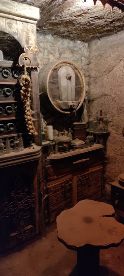 The image shows a cellar or wine storage area in Voskevaz, Armenia. The environment is rustic and appears to be carved into the earth, with rough stone walls and a ceiling covered in what looks like aged wood or rock.
Dominating the center of the frame is a large wooden barrel with metal bands, its bung hole visible. To the left of the barrel, a wooden shelf unit is filled with dark glass bottles, likely wine. A bundle of dried garlic or onions hangs from a decorative wooden carving of an owl or similar creature.
Below the barrel and shelves, a wooden cabinet with multiple drawers and hinged doors is visible. On top of this cabinet and on a small shelf to its right, there are various antique-looking bottles of different shapes and colors, as well as some metal containers and lids that could be part of cooking or storage equipment. One notable object on the shelf is a tall white cylindrical item, possibly a candle or a ceramic vessel.
In the foreground, a unique table with an irregular, organic-shaped top made of a light-colored material sits on a dark base. The floor is concrete or packed earth. The overall lighting is dim, contributing to a sense of age and atmosphere, with cobwebs present on some surfaces, indicating a long-undisturbed storage space. No people are present in the image.