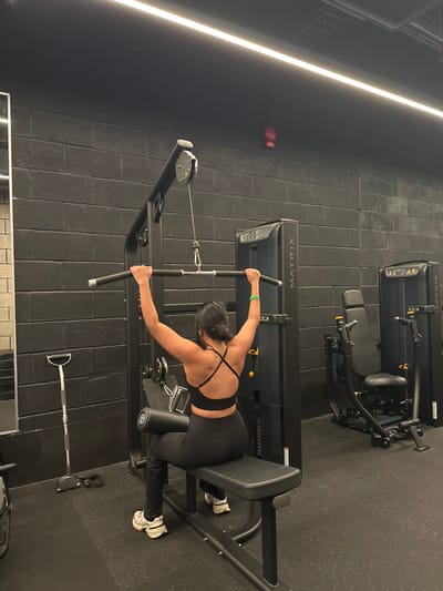 The image depicts an indoor gym facility identified as JD Gyms Dudley, United Kingdom. A female individual is positioned with her back to the camera, seated on a black lat pulldown weight machine, actively engaged in an exercise. She wears a black multi-strap sports bra, black athletic pants, and white athletic shoes with thick soles. A green bracelet is visible on her right wrist. Her arms are extended upwards, gripping a black horizontal pulldown bar attached via a cable and pulley system.
The gym environment features dark grey/black brick walls and a black rubber floor. Overhead, a long, linear LED light strip provides illumination. To the immediate right of the primary subject, another black "MATRIX" branded weight machine, possibly a chest or shoulder press, stands vacant. A third "MATRIX" machine is partially visible further to the right. To the left, a silver T-bar row handle with a black grip is propped against the wall. A distinctive small, red, boxing glove-shaped emergency call point is mounted high on the black brick wall, towards the upper right quadrant of the frame.