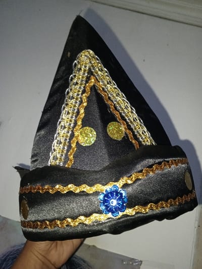 A black, conical fabric headwear, possibly ceremonial, is depicted in Na Sang, Laos. The object features a pointed top and a structured base. Its surface is adorned with gold and silver braided trim arranged vertically on the upper, pointed section. Two circular, glitter-covered gold embellishments are positioned centrally on the pointed area. The base is decorated with a wavy gold trim, and a prominent blue, multifaceted bead arrangement, configured as a floral motif, is affixed to its front. Two small, round, metallic discs are visible on the sides of the base section. A human hand, exhibiting a brown skin tone, is partially visible at the lower left, supporting the headwear from underneath. The background consists of a plain, light-colored wall, illuminated by an unseen light source that casts a subtle shadow behind the object.