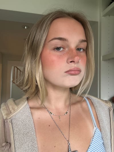 The image presents a close-up, chest-up portrait of a young individual with shoulder-length, straight blonde hair. The person has light skin with visible freckles across the bridge of the nose and cheeks, blue-grey eyes, and a silver hoop nose ring in the left nostril. Prominent rosy-red blush is applied high on the cheekbones, extending towards the nose. The lips are slightly parted, exhibiting a glossy, light pink finish. The individual wears a light blue and white striped garment, likely a bra or bralette, beneath an open, textured light beige or tan hooded zip-up jacket. Multiple silver-toned necklaces are worn: one shorter chain features small beads and a tiny pendant, while a longer chain holds a large, detailed starfish pendant.

The background indicates an indoor residential setting. To the left, a light-colored wall and a partially visible doorway reveal a wooden banister and staircase in the distance. Recessed lighting fixtures are visible in the ceiling. To the right, a light-colored wall features white shelving units with vertical rows of small holes.

There is no discernible information within the image to identify its location as Squamish, Canada, or any other specific geographical area.