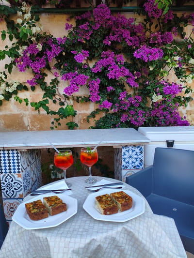 The image shows a table setting outdoors, likely in San Ġwann, Malta. On the table, covered with a light-colored patterned tablecloth, are two white square plates, each holding two slices of what appears to be a savory baked dish, alongside a knife and fork set on a white napkin. Two Aperol Spritz cocktails, complete with straws, garnish, and orange slices, sit behind the plates. The table is positioned in front of a wall covered in lush, vibrant bougainvillea, with purple and white flowers. A small tiled bar or serving area is behind the cocktails to the left. A blue plastic chair is visible on the right side of the frame, suggesting an outdoor dining or relaxation area.