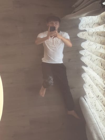 The image, taken in Hanniv, Ukraine, shows a person taking a mirrored selfie. The person is wearing a white t-shirt and dark pants. They are holding a smartphone in their right hand, positioned to capture their reflection. The background is a reflection of what appears to be a room with curtains. The curtains are light-colored with a textured pattern and hang in folds. The floor is a dark, reflective surface, likely a mirror or a highly polished floor, which allows for the reflection of the person and the curtains. The lighting suggests it could be daytime, but the overall tone of the image is somewhat dim. The person's expression is neutral as they focus on taking the photograph. There is no visible text in the image. The scene is intimate and personal, capturing a moment of self-documentation.