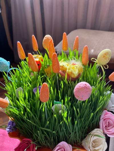 A close-up shot displays an Easter-themed arrangement of green grass, adorned with artificial carrots, roses, and decorative eggs. A pair of small, yellow chicks nestle among the grass blades, surrounded by other colorful Easter decorations. The scene is brightly lit by sunlight. The image provides no discernible location in Aratashen, Armenia.
