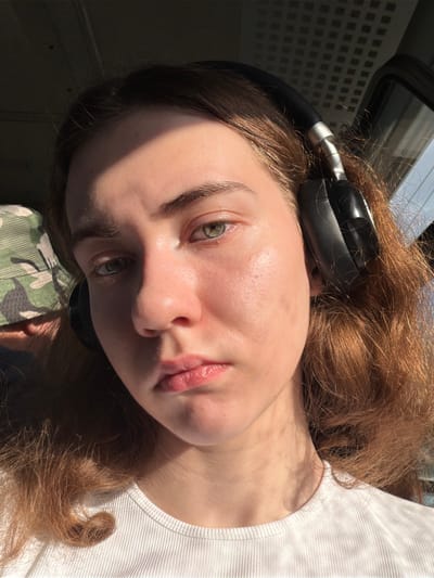 The image is a close-up, frontal selfie of a young person, possibly a teenager, with light brown hair and green eyes. They are wearing black over-ear headphones and a white ribbed t-shirt. The person is looking directly at the camera with a neutral expression. Sunlight is casting a strong shadow across their forehead and right cheek. The background is blurred, but appears to be the interior of a vehicle, possibly a bus or train, with a patterned ceiling visible. A hint of a camouflage-patterned hat or head covering is visible on the left side of the frame. The image was taken in Винзили, Russia.