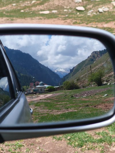 A captivating travel photograph captures a mountainous landscape reflected in a car's side-view mirror, taken in Jeti-Ögüz, Kyrgyzstan. The primary subject is the reflection of a deep valley, flanked by steep slopes covered in dense evergreen forests. In the distance, majestic snow-capped peaks rise under a partly cloudy sky with patches of blue.
In the mid-ground of the reflection, a small settlement is visible, featuring a distinctive building resembling a mosque with light blue-green domes and a minaret. A dirt road or path winds through the valley floor, passing by this structure and other smaller buildings. Green grass, bare earth, and scattered rocks make up the foreground of the reflected landscape, with a few wooden utility poles hinting at infrastructure.
The left side of the original image shows the grey metallic body of the car, likely the driver's or passenger's door, with subtle reflections on its surface. The black frame of the side-view mirror sharply defines the reflected scene. Outside the mirror, in the immediate foreground, is unpaved ground with green grass and light-colored stones, suggesting the car is parked or moving slowly on a rural track. The time of day appears to be daytime, with clear visibility of the distant mountains. No discernible text is present in the image.