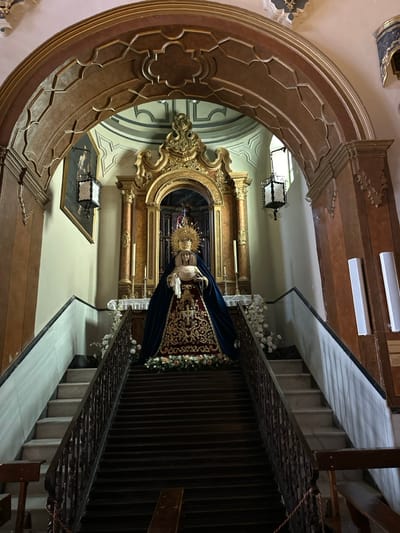 The interior of the Santuario de María Auxiliadora (San Roque) in Ronda, Spain, features a prominent religious display. A sorrowful statue of the Virgin Mary, known as a Mater Dolorosa, is centrally positioned at the top of a wide, dark wooden staircase. The statue is adorned with a golden, radiating halo, a dark blue velvet cloak richly embroidered with gold thread, and an intricately patterned underskirt. Her hands are clasped, holding a white cloth, and a rosary hangs from her right hand.

The statue stands within an ornate, gilded wooden reredos featuring fluted columns and elaborate carvings, set against a dark background in its central niche. Two tall golden candlesticks with lit candles flank the statue on the altar. The base of the statue, at the top of the steps, is decorated with an arrangement of white and light pink flowers interspersed with greenery.

The central wooden staircase, comprising approximately 15 steps visible, has dark banisters and leads directly to the statue. This central staircase is flanked on both sides by narrower, lighter-colored marble staircases with dark handrails, ascending to the same elevated platform as the altar. The entire religious ensemble is framed by a large, intricately carved wooden archway with geometric and quatrefoil patterns.

The surrounding walls are light-colored. A dark framed painting hangs on the left wall, below a suspended ornate dark metal lantern with glass panels. A similar lantern is visible on the right side. On the right wall, two vertical, rectangular white light fixtures are mounted. A partial view of a dark wooden pew is visible in the lower right foreground. No individuals are present in the scene.