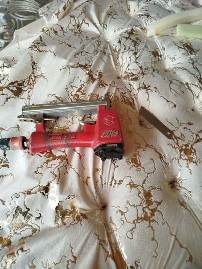 A partially upholstered item, possibly a headboard or cushion, sits on a patterned white and gold fabric. Depressions suggest tufting. A red pneumatic staple gun rests on the fabric, near a small rectangular piece of metal. Some foam pieces are visible in the upper right. The scene implies ongoing upholstery work.