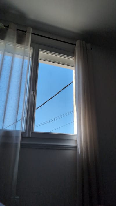 A view through a window in Cholet, France, reveals a bright blue sky with a few thin, dark wires crossing it. The window is framed by sheer white curtains that are partially drawn, allowing sunlight to filter through. The room itself is dimly lit, with shadows cast on the wall beneath the window. The overall impression is one of a quiet, domestic scene with a glimpse of the outside world.