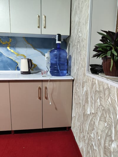 The image shows a close-up view of a kitchen counter area, likely located in a residential setting in Navruz, Uzbekistan. A large blue water cooler jug with a black electric pump on top is prominently featured. To the left of the water cooler is a beige and brown electric kettle with a diamond pattern. A small, clear glass with some liquid and a red design is placed next to the kettle. The counter itself has a marble-like pattern.
Above the counter, there are two beige cabinet doors with gold handles. The backsplash behind the counter is a decorative blue and gold marbled pattern.
To the right of the water cooler, the wall is covered in wallpaper with a repeating feather or leaf pattern in shades of beige and white. A potted plant with dark green and purple leaves sits on a shelf to the right, next to a black plastic bag and a white plate. The floor in front of the counter is covered with a red carpet. A white cable is visible running from the water cooler pump, draped down towards the counter. The overall scene suggests a functional, domestic space.