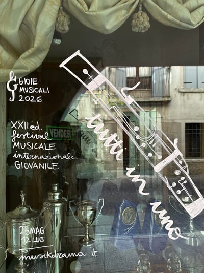 The image is a photograph taken at street level, looking through a shop window in Maser, Italy. White chalk art is drawn on the glass, depicting a musical instrument resembling a bassoon, along with text. To the left of the instrument drawing, text reads "GIOIE MUSICALI 2026" with a musical G clef. Below this, it says "XXII ed. festival MUSICALE internazionale GIOVANILE". A green sign in the background reads "VENDESI" (for sale).
Further down, on the left side of the window, a silver trophy is visible, with text written on it in chalk: "25MAG 12 LUG". To the right of this trophy is a silver shaker and another silver trophy. Behind these objects, two blue awards with gold medallions are displayed. In the bottom right corner, the text "musicheilrama.it" is written on the glass.
The shop window reflects the street outside, showing a building with balconies and windows, suggesting an urban or town setting. The overall lighting indicates it is daytime, with some natural light filtering through the window. The chalk art suggests the shop may be related to music or events.