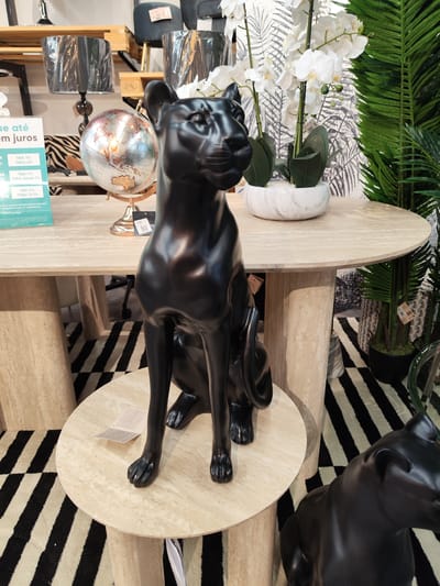 This image captures an indoor retail display, likely within a home decor or furniture store, in Ponta Delgada, Portugal. The central subject is a sleek, glossy black panther statue, depicted in a sitting position on a small, round, light-colored table with a pedestal base. Another similar black panther statue is partially visible in the bottom right corner.
Behind the prominent panther, a larger, round, light-colored table with a matching pedestal base serves as a display surface. On this table, a vintage-style globe on a metallic stand is visible, alongside a table lamp with a dark fabric shade and an ornate base. To the right, a white artificial orchid plant in a marble-patterned pot adds a touch of greenery, complemented by several large artificial palm fronds arranged around the back of the table.
The background includes shelving units with various items, notably a dark grey upholstered stool and a wooden pallet marked "EPAL". The floor is covered with a striking black and white striped rug or carpet, creating a modern and graphic contrast.
Visible text in Portuguese from a sign on the left advertises financing options: "ue até em juros" (up to in interest), "TAN: 0% TAEG: 0%", "TAN: 0% TAEG: desde 3%", and "TAN: 0% TAEG: 10,1%", followed by details about "comissão de formalização" (formalization commission) and "mensalidade" (monthly payment). A price tag reading "22€" is visible near the globe and also on a tag near the main panther statue. Another tag displays "LIVING". No people are present in the scene. The overall aesthetic is contemporary and elegant, featuring a neutral color palette with bold accents.