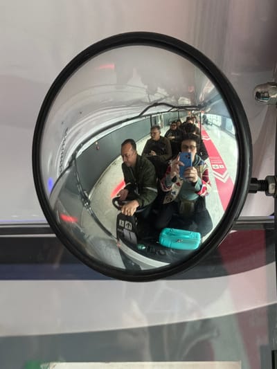 The image shows a reflection in a convex mirror of a group of people seated in a track-based vehicle. The vehicle appears to be in motion, possibly on a ride or transport system. The people are facing forward and are captured from the chest up. In the foreground of the reflection, a person is holding up a blue smartphone and taking a selfie. This person is wearing glasses and a colorful jacket. A teal-colored case is visible on the floor of the vehicle in front of them. The environment reflected in the mirror appears to be an enclosed, dimly lit space with red and white markings on the floor, suggesting a pathway or track. The overall scene suggests a recreational or entertainment setting. This image was taken at the Guarulhos International Airport (GRU) in Guarulhos, Brazil.
