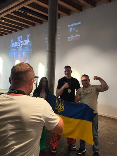 The image depicts an indoor event, likely a sporting competition, in Romanshorn, Switzerland. In the foreground, a person from behind is wearing a white t-shirt and a camera strap. To the right, three individuals are standing together. The two men in the center and right are holding a blue and yellow Ukrainian flag. The man on the left, wearing a black t-shirt and a gold medal, is posing with a closed fist. The man on the right, in a tan t-shirt, is flexing his bicep. A woman in a green garment is standing behind and to the left of the men. Behind them, a large projector screen displays event information, including the title "BABAEV'S CUP" and the date "JUNE 13TH 2026", along with the address "KORNHAUS, FRIEDRICHSHAFENSTRASSE 54, 8590 ROMANSHORN, SWITZERLAND". The environment appears to be a hall with wooden beams on the ceiling and a concrete pillar.