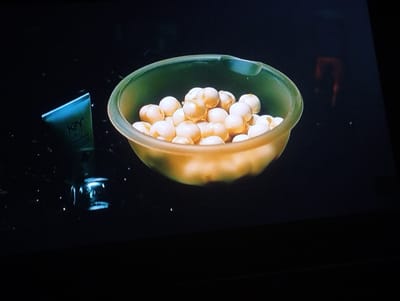 A bowl of tan-colored spherical items is centered on a dark surface. To the left of the bowl is a tube of lubricant. The overall lighting is dim. The location is undetermined. No people are visible.