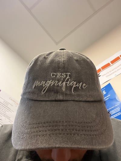 The image displays an upward-angled view of an individual partially obscured by a dark grey, distressed baseball cap. The cap features light-colored embroidered text on the front stating "C'EST magnifique." Only the lower portion of the individual's nose and chin, consistent with a person wearing a beard or heavy stubble, are visible beneath the cap's brim. The individual is wearing a dark grey garment that covers the shoulders.
The background reveals an indoor environment with light-colored walls and a white ceiling composed of rectangular panels. On the left side, a white document, likely a "Certificat," is partially visible, displaying text including "ISO 9001" and "DR REVULUS." On the right, an orange and white banner or poster reads "SALLE D'ATTENTE" (Waiting Room) and "CHAMPS OUVERT." Below this, a blue document or poster is visible, containing text such as "SUR" and explicitly stating "Stains," followed by a phone number "01-49-71-45-70."
The scene is located within a building in the city of Stains, France, as confirmed by the legible text on the blue poster.
