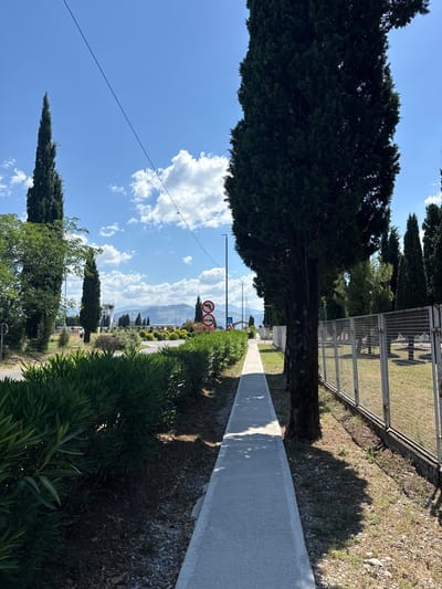 The image depicts a pedestrian walkway alongside a road, bordered by dense green bushes on the left and a metal fence with a grassy area and trees beyond on the right. Tall, slender cypress trees are prominent on the left side of the walkway, with a larger, darker tree trunk in the foreground on the right. In the distance, an airport control tower is visible, surrounded by buildings and more trees, with mountains forming the backdrop under a bright blue sky with scattered clouds. Traffic signs, including one indicating no entry and another for a turn, are present along the road. The overall environment suggests an outdoor setting, possibly near an airport or a thoroughfare in Golubovci, Montenegro.
