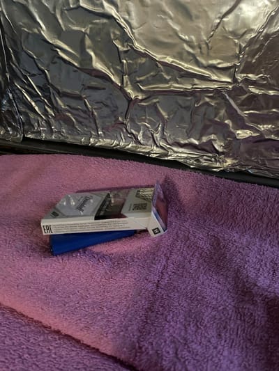 The image shows a close-up view of a pack of Davidoff cigarettes lying on a purple, fuzzy surface, possibly a blanket or towel. The cigarette pack is white with some text and graphics, including a barcode and the "EAC" certification mark. The brand name "Davidoff" is prominently displayed. Beneath the cigarette pack, a portion of a blue object, likely a lighter, is visible. The background features a crinkled, metallic silver material, suggesting a foil-like surface that is reflecting light. The scene is indoors, and the lighting appears to be artificial, casting shadows and highlights on the metallic background. There are no people or identifiable landmarks of Yokohama, Japan, present in the image.