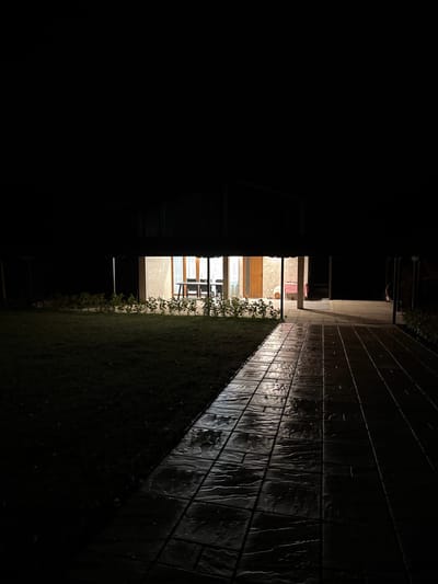 This nighttime image captures an outdoor area leading to a building in Peyovstsi, Bulgaria. The foreground consists of a dark, low-cut grassy lawn on the left and a wet, dark gray paved walkway composed of rectangular, textured tiles on the right, which reflects the internal light from the building. A low line of green foliage separates the lawn from the building's facade. The midground features a building with light-colored walls and large glass doors or windows, through which interior artificial light is prominently visible. Inside the illuminated section, a rectangular table is discernibly set with multiple dark chairs around it. To the right of this area, another illuminated interior space or doorway is partially visible, showing a warmer light source. The upper portion of the image, the background beyond the illuminated structure, and the immediate foreground of the lawn are enveloped in darkness. No individuals or discernible actions are present in the scene.