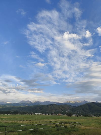 The image depicts a serene landscape in Қырғауылды, Kazakhstan. The scene is dominated by a vast blue sky adorned with wispy white clouds, suggesting a pleasant, possibly late afternoon or early evening atmosphere. Below the sky, a range of majestic, green mountains stretches across the horizon, their peaks catching the light and hinting at snow. Nestled in the foothills are scattered houses and buildings, forming a small, tranquil village. In the foreground, a grassy field is marked by a black and white striped fence, adding a touch of man-made structure to the natural beauty. The overall mood is peaceful and expansive, capturing the quiet grandeur of the Kazakh countryside. There is no visible text in the image.