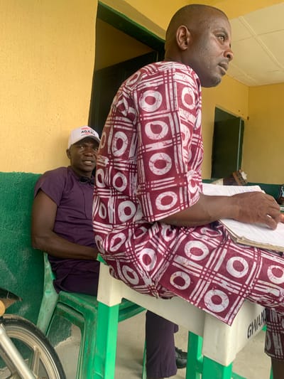 The image shows two men in Tabu, Nigeria. One man, dressed in a maroon and white patterned shirt, sits on a green plastic chair with a white top. He holds a notepad. He is looking up. Below him, another man in a purple shirt and a white "AGARA" cap sits on a green plastic chair with a white top and looks up at the man above. The scene takes place outside a yellow building with green trim. A bicycle wheel is visible to the right.