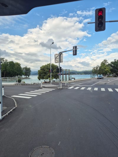 The image shows an intersection in Seewalchen am Attersee, Austria, on a cloudy day. A traffic light in the upper right corner is red. A pedestrian crossing is visible in the foreground, and the Attersee lake is in the background, with trees and hills on the opposite shore. A dark-colored car is driving away from the camera on the right side of the road. The road surface appears to be asphalt, with white markings for the crosswalk and lane dividers. A streetlamp is visible on the left side of the intersection.